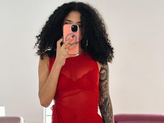 Lanna Jhonson, met krullend zwart haar, draagt een doorschijnende rode jurk en houdt een roze telefoon vast met een ringhouder, waarop een tatoeage te zien is. arm. Helder witte achtergrond, waarop een aantrekkelijke selfie is te zien..