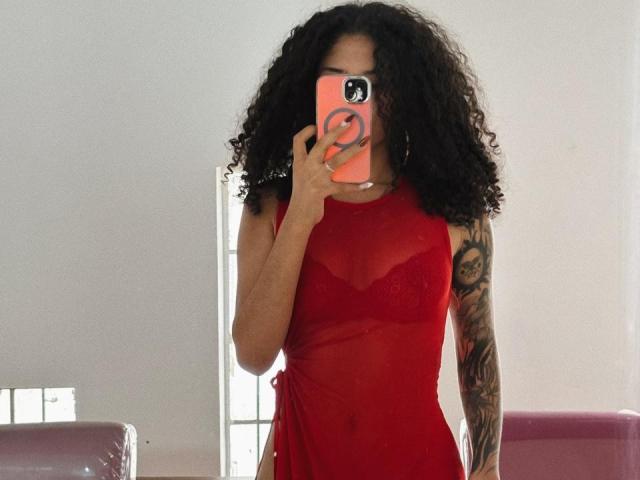 Lanna Jhonson está em um quarto bem iluminado, vestindo um vestido vermelho transparente que revela seus mamilos. Ela segura um telefone vermelho com um adesivo de rosto sorridente, cobrindo seu rosto. Braço com tatuagem visível..
