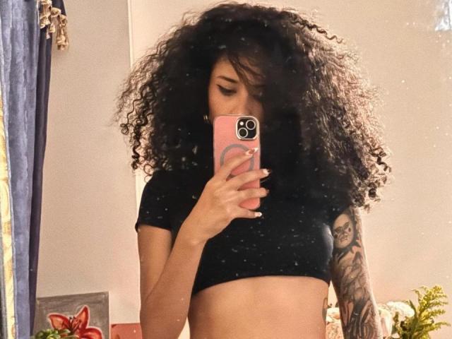 Fotografia de Lanna Jhonson, tirando uma selfie no espelho com um celular rosa. Ela tem cabelo preto encaracolado, uma blusa cropped preta e um braço tatuado. Seus seios estão parcialmente visíveis. Incrivelmente sexy..