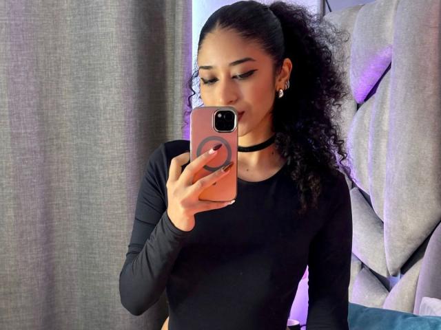 Lanna Jhonson, eine attraktive Frau mit lockigem, schwarzem Haar, macht ein Selfie vor einem Spiegel und trägt dabei ein schwarzes Oberteil mit langen Ärmeln. Eine Frau mit einem schwarzen Kragen, die ein orangefarbenes Telefon hält, mit einem gesteppten Kopfteil im Hintergrund..