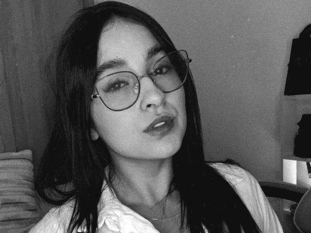 Black and white photo of EmmaWaltter with long dark hair, wearing glasses and a white shirt. She has a sultry expression, with slightly parted lips. Her eyes are half-lidded.