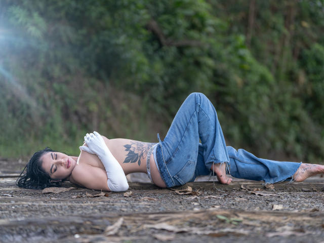 Antonella Ferrer è sdraiata su un sentiero sterrato, con le gambe piegate, indossa jeans blu e una camicetta bianca in pizzo, che rivela un grande tatuaggio sulla sua pelle. lato. I suoi capelli neri sono spettinati, gli occhi chiusi. Incredibilmente sexy..