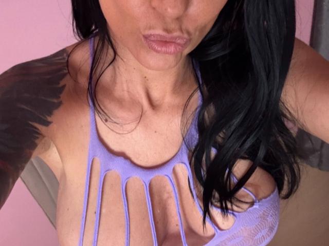 Close-up photo of AndraD with long black hair, pouty lips, and a tight, white, cut-out tank top showing large breasts and cleavage. Sexual tension, seductive pose, visible cleavage.