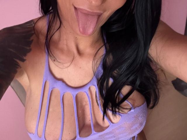 Close-up photo of AndraD with black hair, sticking out tongue, wearing a sheer white top with large cutouts revealing large breasts. Background is pink, tattoo on shoulder visible. Sexual tension.