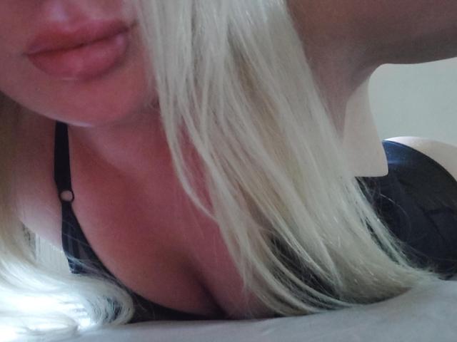 Close-up photo of YourCoronet lying on their stomach, blonde hair partially covering face, pink lips slightly parted, black bra straps visible, cleavage emphasized, bright light in background.