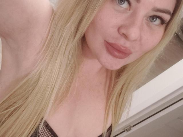 Close-up selfie van YourCoronet met lang blond haar, blauwe ogen en volle lippen. Een zwarte kousenbroek aan, voorovergebogen, decolleté zichtbaar. De huid is licht, de uitdrukking is licht verleidelijk..