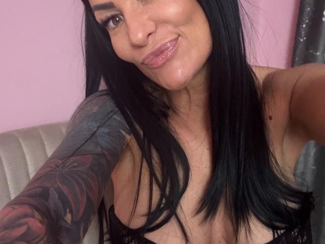 Close-up of AndraD, a tattooed woman with long black hair, glossy pink lips, and a cleavage reveal. She's smiling slightly, glistening lips suggest wetness. Foreground focus, intimate selfie vibe.