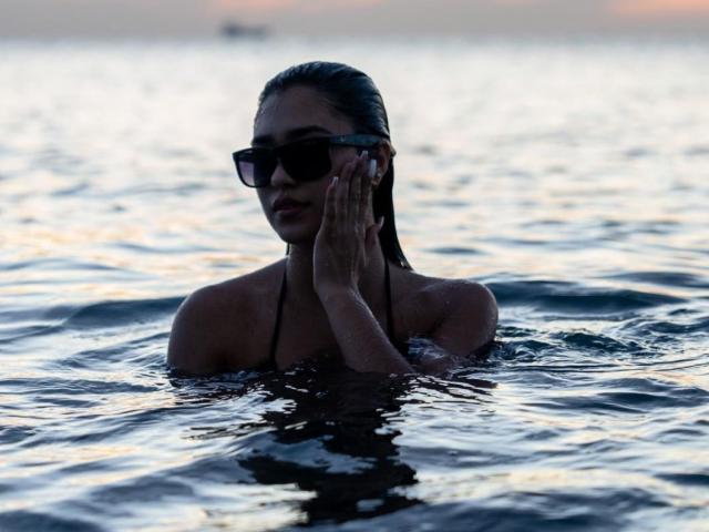 Fotografía en silueta de MiaaFerrer, con el pecho sumergido en el agua, las manos juntas frente al rostro, y usando gafas de sol oscuras. Iluminación por la luz del atardecer, agua reflectante, sutil tensión erótica, expresión seria, cabello oscuro peinado hacia atrás..