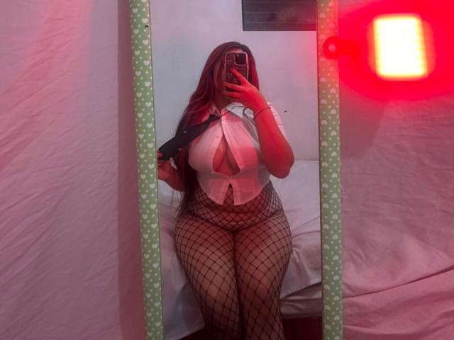 SeleneVladis scatta un selfie allo specchio, tenendo in mano un telefono con il flash rosso. Indossa collant a rete e una maglia bianca con scollatura profonda, che mette in risalto il suo seno generoso e i suoi fianchi sinuosi..