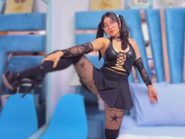 Genesiss strikes a provocative pose, one leg raised high, wearing a black lace-up top, fishnet arm sleeves, and mini skirt. Black thigh-high boots, red lipstick, blue background. Sexual tension.