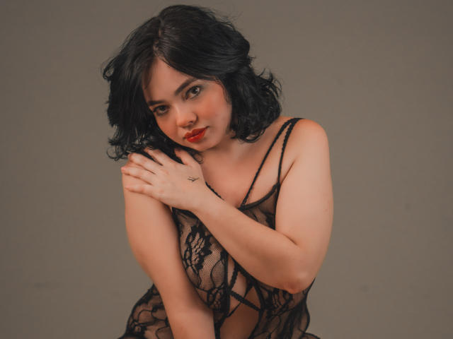 AprilSwett, con i capelli neri e ondulati e il rossetto rosso, indossa una trasparente camicetta di pizzo nero che mette in mostra il suo décolleté. È china in avanti, con le braccia incrociate sul petto, assumendo una posa seducente..