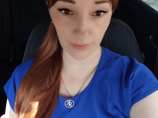 Fotografía de primer plano de MissieLorrie, de piel clara y cabello castaño recogido en una coleta, vistiendo una camisa azul y un collar de plata. con un símbolo de paz, sentada en un coche, atractiva, mirada directa..