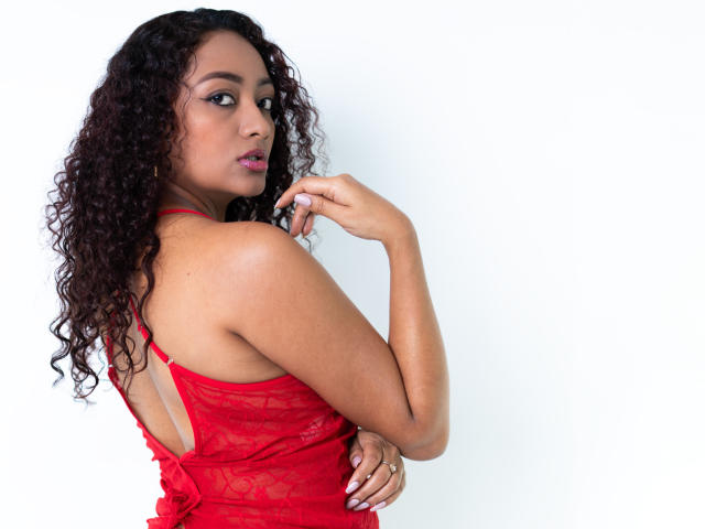 LaurenHotestx, com cabelos pretos e cacheados, veste um vestido vermelho de renda e olha por cima do ombro com uma expressão sedutora. A mão dela toca o queixo, realçando sua figura curvilínea e seus seios grandes..