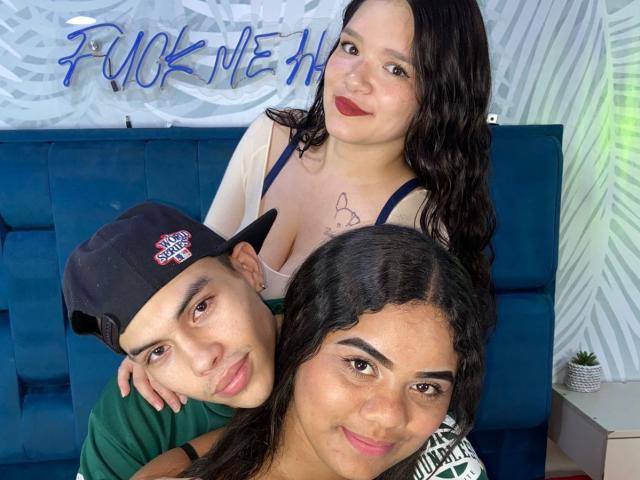 Fotografia di TripleTemptation e di due uomini, primo piano. TripleTemptation con capelli neri, rossetto rosso, scollatura e un tatuaggio, è china in avanti. Un uomo con una camicia verde e un cappello nero, l'altro in primo piano. Un neon con la scritta "Fuck Me"..