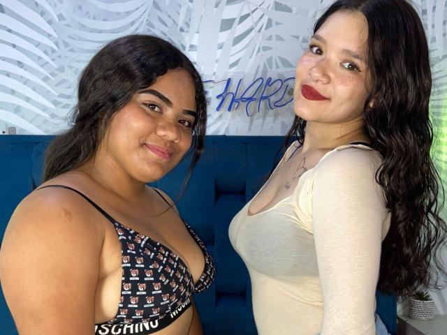 Due donne, TripleTemptation, che stanno vicine l'una all'altra. A sinistra, una indossa un reggiseno senza ferretto nero con motivi, a destra una maglia a maniche lunghe bianca, entrambe con capelli scuri e che sorridono. Illuminazione intensa, posa informale, leggera tensione sessuale.