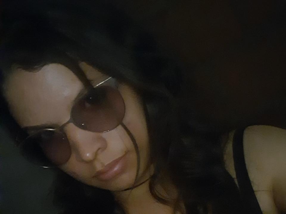 Close-up de LolliMoon com cabelo cacheado escuro, usando óculos de sol marrons grandes e uma camiseta preta, inclinada para frente com um um sorriso discreto e sedutor. A iluminação baixa realça o seu lado sensual..