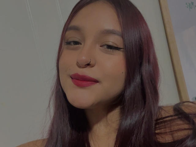 Close-up of MarianaVargasX, a young woman with long, straight black hair, red lipstick, and a nose ring, smiling subtly. Soft lighting, slight shadow on right side. Fucking sexy.