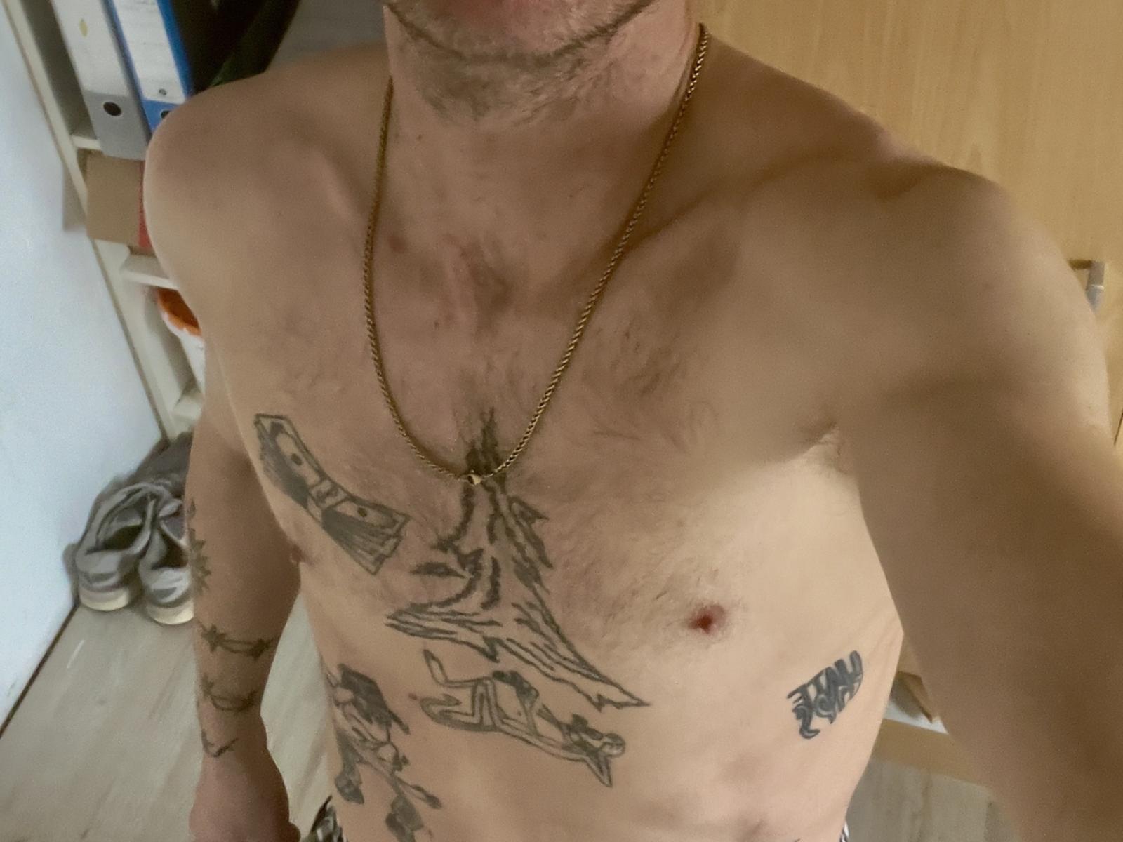 Man zonder shirt, FourTwwo, met tatoeages op de borst en armen, draagt een gouden ketting, staat met een licht voorovergebogen houding. met een gespierd, behaard bovenlichaam, in een rommelige kamer.