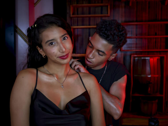 RickeyNgySweet with long dark hair and red lipstick, wearing a black spaghetti strap top, stands close to a muscular man with short curly hair, who touches her shoulder. Red lighting.