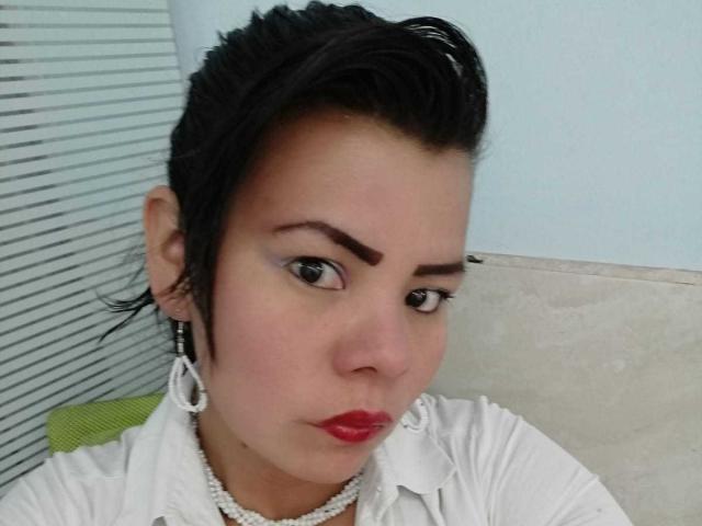 Primo piano di KiaraXMoon, una donna latina con capelli neri corti, rossetto rosso e camicia bianca. Indossa una collana di perle e grandi orecchini a cerchio. La sua espressione è leggermente sensuale. Molto attraente..