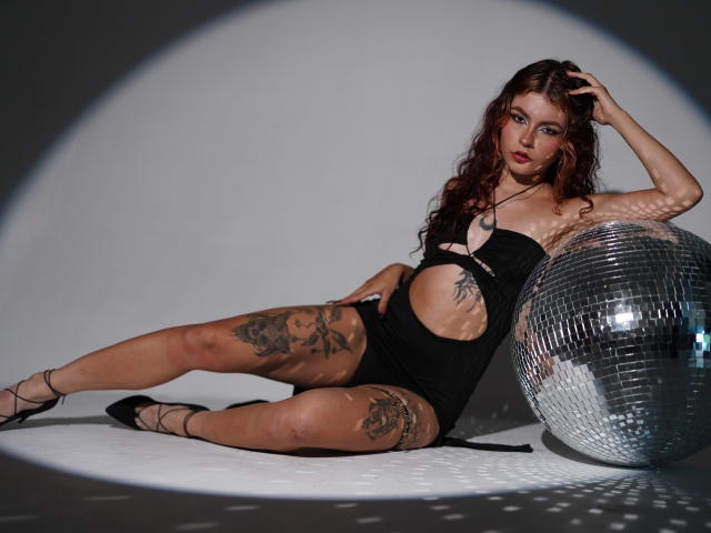 ScarletBlazeX lehnt sich an einen glänzenden, silbernen Discokugel, trägt einen schwarzen, figurbetonten Body mit Ausschnitten, der Tattoos an ihrem Oberschenkel freilegt. und modern. Sie blickt verführerisch, die Beine gespreizt, und erzeugt dadurch eine intensive sexuelle Spannung..