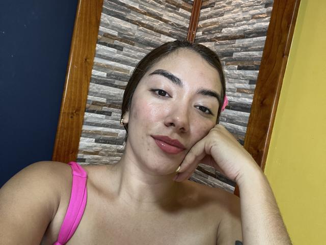 SharonKlart, close-up selfie, topless with pink strap, resting head on hand, dark hair pulled back, light skin, slight smirk, wooden shutter background, yellow wall, shiny skin, relaxed pose, slight sexual tension.