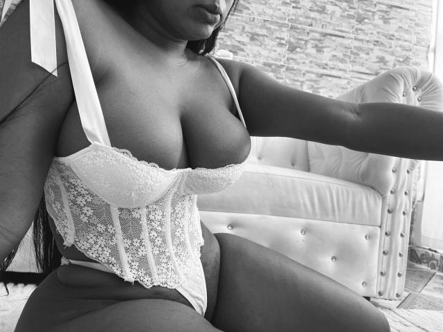 Fotografía en blanco y negro de DiosaEbano, de piel oscura, vestida con un corpiño de encaje con grandes senos, sentada en un sillón de cuero con capitonado. silla. Su brazo derecho se extiende hacia adelante, creando una pose seductora..