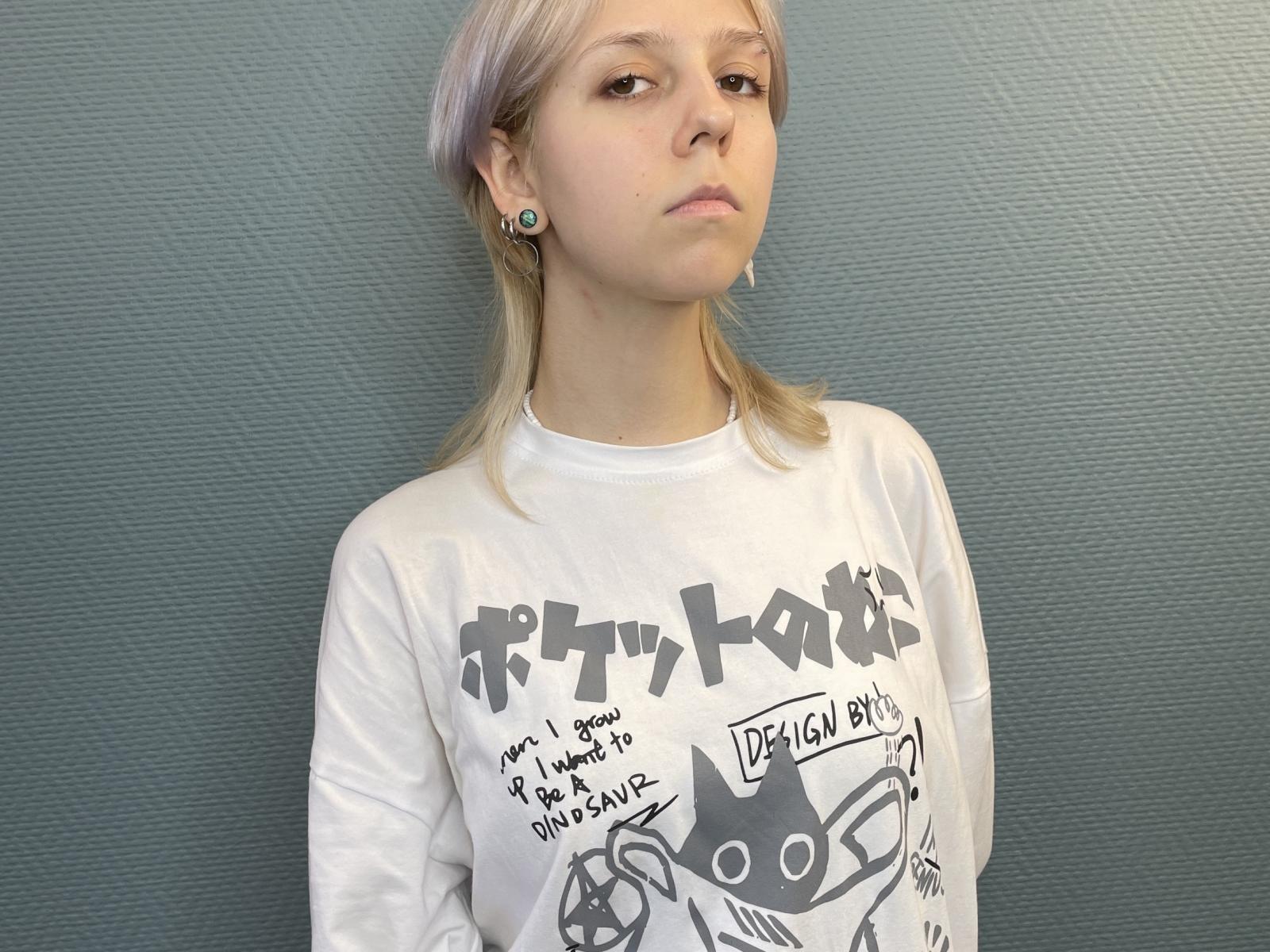 Fotografia di KateIbey, una giovane donna con capelli biondo chiaro, che indossa una t-shirt bianca con una grafica, in piedi di fronte a uno sfondo strutturato. Parete di colore blu-grigio, che guarda leggermente a sinistra, con un'espressione seria. Illuminazione mirata.