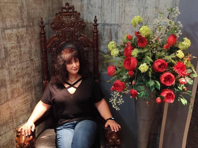 JannaCurvy, sentada em um trono de madeira escura, vestindo uma blusa preta com alças cruzadas e jeans azuis, ao lado de um vaso de rosas vermelhas e amarelas vibrantes. Absolutamente deslumbrante..