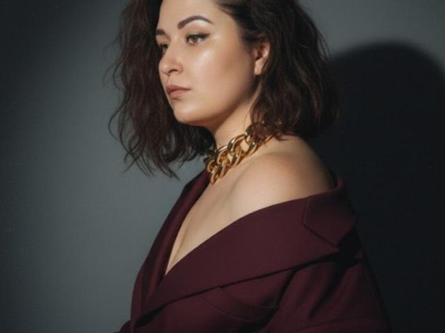 Photograph of JannaCurvy, facing left, with dark wavy hair, brown off-shoulder top, gold chain necklace, slight smirk, shadowed background. Her gaze is confident, sexual tension evident in her expression.