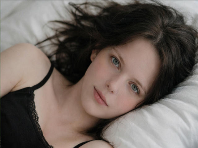 Gros plan de DannaSt allongée sur un oreiller blanc, ses cheveux bruns foncés éparpillés, portant un caraco en dentelle noire, peau pâle, yeux bleus. yeux, légère rougeur, regard séduisant, sourire discret, éclairage doux.