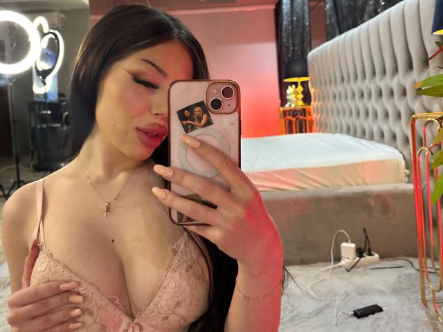 AlessiaNoire, una mujer asiática con cabello largo y negro, se toma un selfie frente a un espejo en un dormitorio. Ella lleva un sujetador de encaje rosa que realza sus grandes senos, y sostiene su teléfono con una mano. El dormitorio cuenta con un cabecero acolchado e iluminación suave..