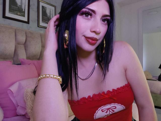 IsabellaaRossi, dark-haired woman with red lipstick, wearing a red strapless top with a bow, gold bracelet and earrings, hand in hair, looking at camera, sexy, confident, bedroom setting.