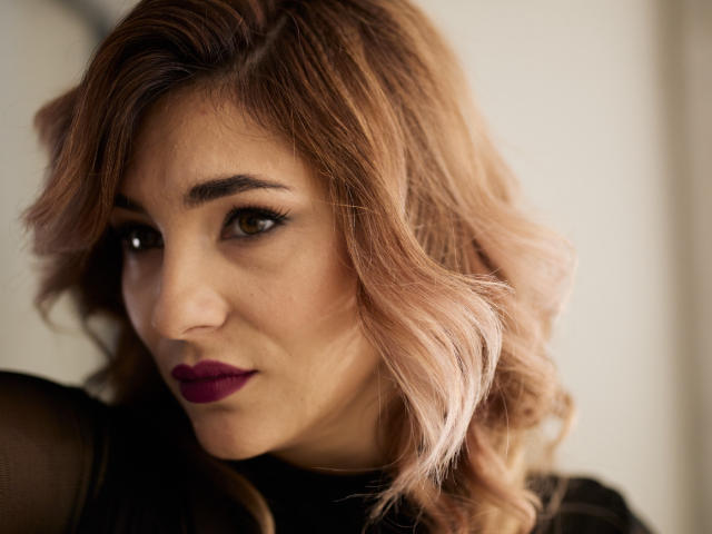 Close-up van DakotaBey met halflang, golvend, lichtbruin haar, donkere eyeliner en donkerrode lippenstift. Zacht licht accentueert haar gezicht, en werpt schaduwen op haar linkerwang. Subtiele, verleidelijke uitdrukking.