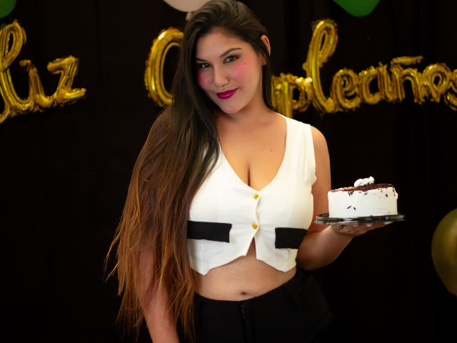 Maria Rowe, eine kurvige Frau mit langen braunen Haaren, die ein weißes Crop-Top mit schwarzen Streifen trägt, hält ein Stück Kuchen in ihrer Hand. Rechte Hand. Sie lächelt, mit rosa Lippenstift..