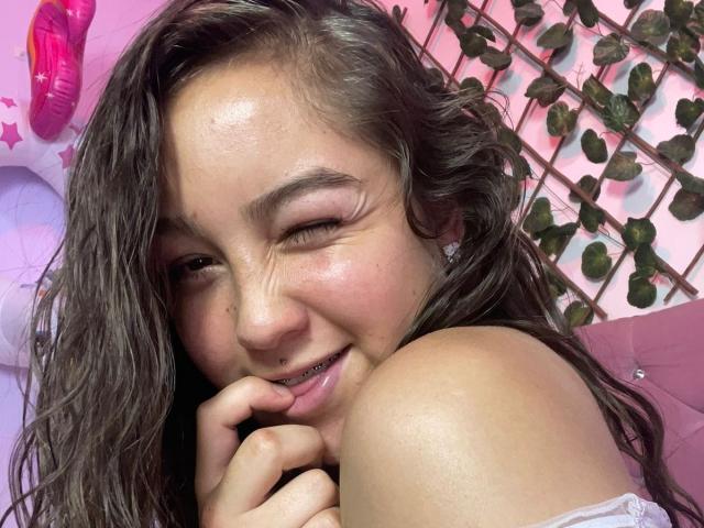 Close-up de EmmaQuintana, uma mulher de cabelos escuros e ondulados, com o cabelo molhado, sorrindo com os olhos fechados, mordendo o dedo, que está rosado. blush, blusa branca com um ombro descoberto, fundo com folhagem, brinquedo sexual vermelho à esquerda. Muito quente..