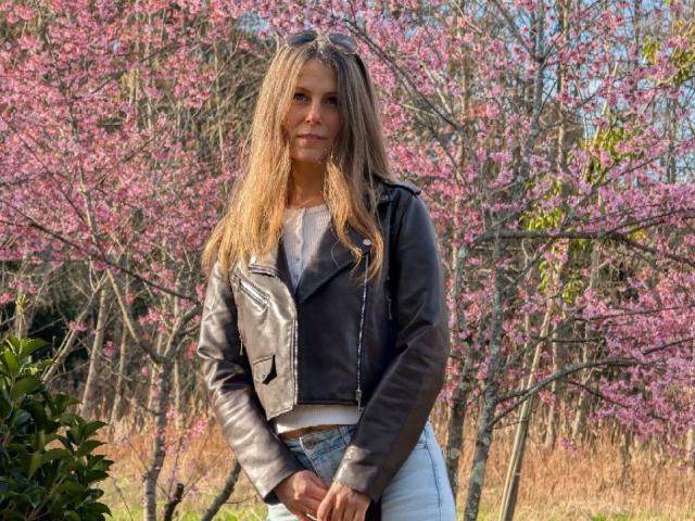 AuroraStar, con i suoi lunghi capelli biondi e umidi, è in piedi di fronte a fiori di ciliegio rosa, indossando una giacca di pelle nera e un jeans. Lei guarda direttamente la telecamera con un'espressione seria. La luce del sole illumina i suoi capelli bagnati e la giacca..