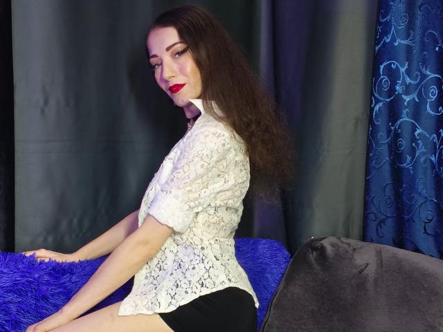 Silerta with long, dark curly hair, red lipstick, and white lace blouse, leaning forward on a blue textured surface, black pants, grey and blue patterned curtains in the background. Fucking sexy.