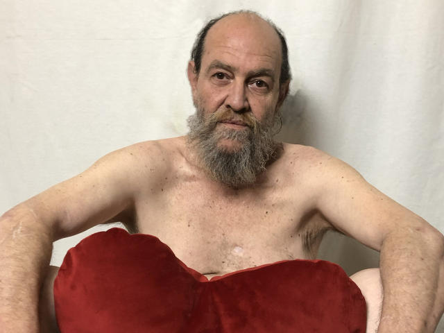 Homme d'âge moyen, chauve sur le dessus et avec une barbe grise, torse nu, tenant un grand oreiller en velours rouge devant son entrejambe, regardant. Directement face à la caméra, avec un sourire narquois. Tension sexuelle.