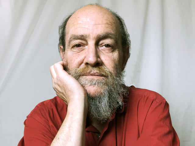 UmbertoHottie, um homem de meia-idade com a cabeça careca e barba grisalha, veste uma camisa vermelha e apoia a mão na bochecha. Sua expressão é ligeiramente sugestiva, com um toque de sorriso..