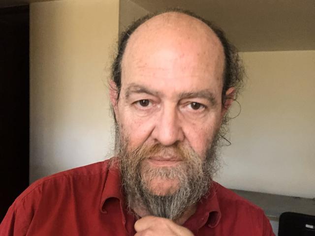 UmbertoHottie, um homem de meia-idade com a cabeça careca e uma barba grisalha, veste uma camisa vermelha. Ele está segurando o queixo com um sorriso discreto, transmitindo uma sensualidade casual e sem barba..