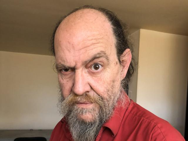 UmbertoHottie a une tête dégarnie, une barbe épaisse et grise, et des yeux intenses et perçants. Portant une chemise rouge, il fixe l'appareil photo avec un regard intense, la tête légèrement inclinée. L'éclairage met en valeur son visage..