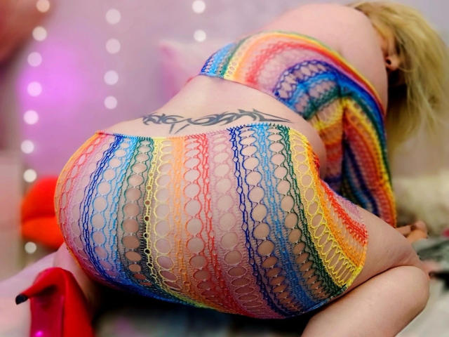 HotSandy4You- seksueel, knielend, met de billen naar de camera gericht, draagt regenboogvisnetlingerie, zichtbare tatoeage op de onderrug, blond. haar, rode hoge hakken, wazige lichtjes op de achtergrond, kleurrijk, sexy, uitdagende pose.