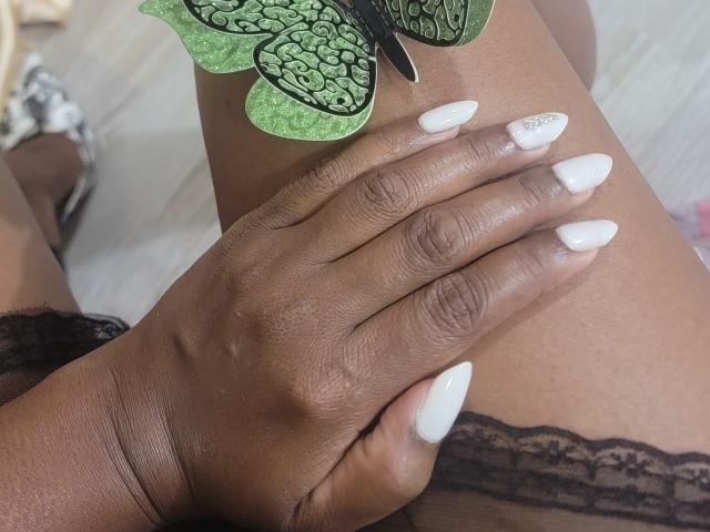 Close-up de uma mão de pele escura com unhas brancas e brilhantes, apoiada na cintura de uma mulher, coberta por renda preta. Um adesivo com formato de folha verde cobre parcialmente a parte superior da coxa dela. Tensão sexual, lingerie..