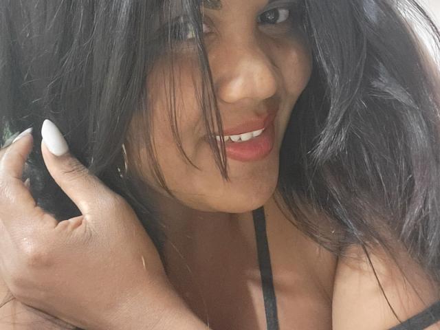Close-up photo of FelyLove, a dark-skinned woman with long black hair, smiling with red lips, white nail polish, and a black strap dress. Fingering her hair, showing sexual tension.
