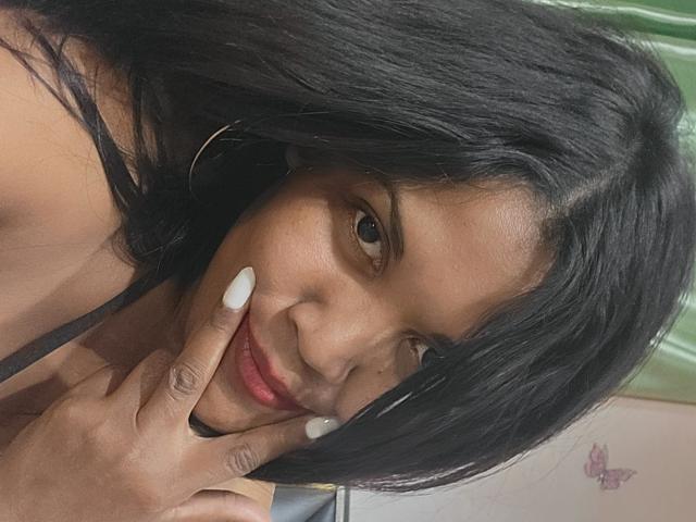 Close-up photo of FelyLove with dark brown skin, black hair, and white nails, making a playful face while touching her cheek with her fingers. Visible red lipstick, green background.