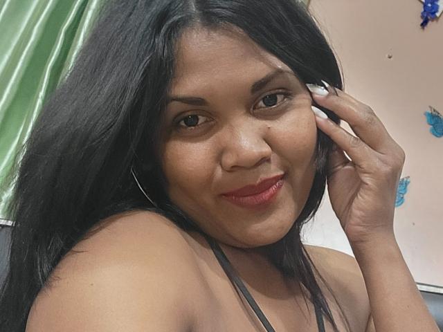 Close-up of FelyLove, a curvy woman with long black hair, red lipstick, and brown eyes, touching her face with one hand, wearing a black strap top, slight smile, green curtain background.