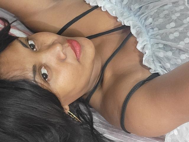 Close-up of FelyLove, lying down, wearing a white lace top with black straps, red lipstick, dark hair, and a black choker, looking seductively at the camera, hand on face.