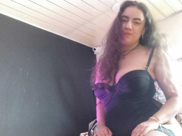 MiaBrooklyn, met lang, krullend bruin haar, draagt een strakke zwarte jurk zonder bandjes die haar grote borsten accentueert. uitsnijding. Ze kijkt met een licht opgewonden uitdrukking in de camera. Slechts zwak verlichte kamer.