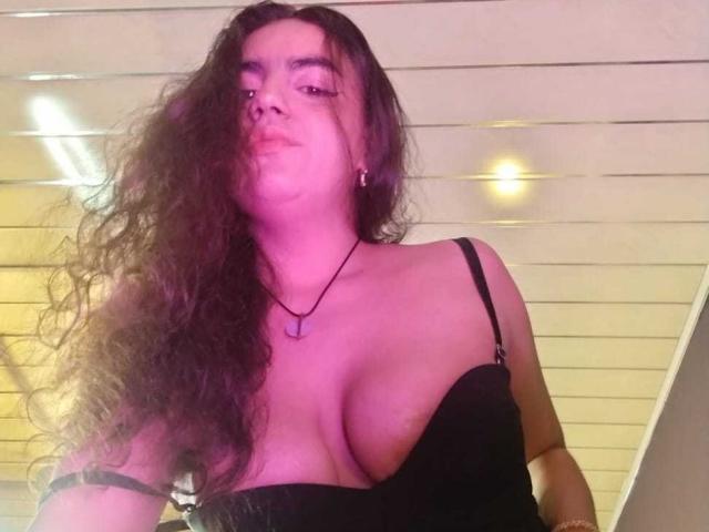 Photograph of MiaBrooklyn, a curvy woman with long, wavy brown hair, wearing a black strapless top that reveals her large breasts. Her expression is confident, with pink lighting.